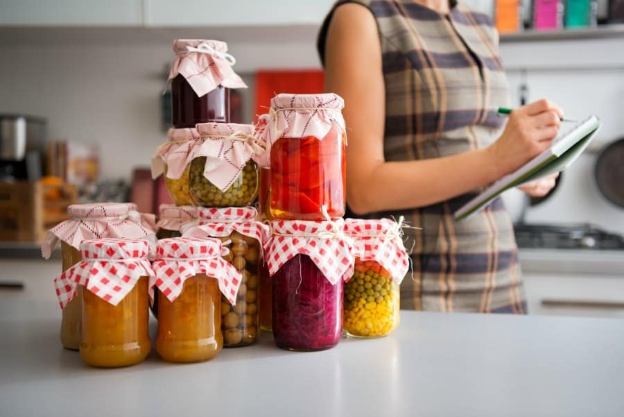 Preserved homegrown food in canning jars on kitchen counter, woman in the background making notes